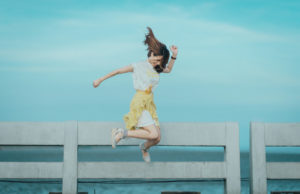 Brunette woman in a white and yellow summer outfit jumps happily on a pier by the ocean