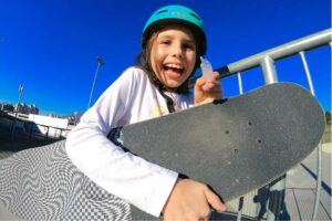 young girl wearing a helmet, skateboard under her arm, smiles and holds up her mouthguard